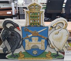 A wooden coat of arms showing a white swan and a black swan facing each other. A sheild showing a castle and cross swords is betwwen them, headed by a portcullis infront of a tree, with a crown on top.